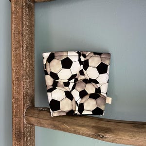 May include: A fabric coffee cup sleeve featuring a repeating pattern of black and white soccer balls. The sleeve is rectangular and has a small, neutral-colored tag on the side. It rests on a wooden ladder rung against a light blue wall.