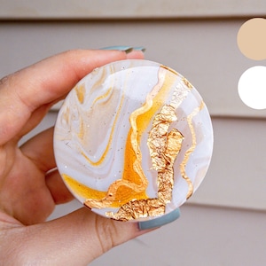 Puede incluir: Un objeto decorativo redondo, blanco y dorado, con detalles de pan de oro. El objeto tiene un efecto marmolado con remolinos dorados y blancos. El objeto está sostenido en una mano.
