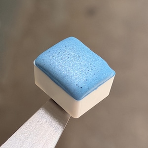May include: A small, square watercolor paint pan with a white plastic base and a textured, shimmering light blue pigment block filled to the top. The paint has a slightly porous, granular surface appearance and is held by metal tweezers.