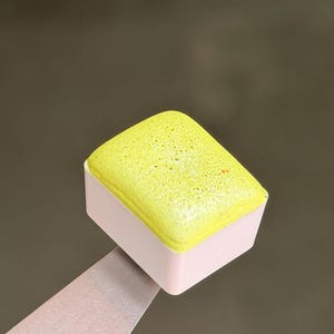 May include: A rectangular watercolor paint pan with a light pink plastic base and a textured, vibrant yellow pigment block on top. The paint surface shows a porous, granular consistency with a small orange speck visible on the upper right corner.