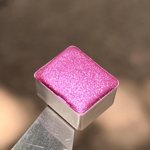 May include: A rectangular watercolor half pan filled with a vibrant, glittery magenta pigment. The paint has a textured, sparkling surface and is contained within a white plastic casing, held by a metallic tool at the bottom corner.