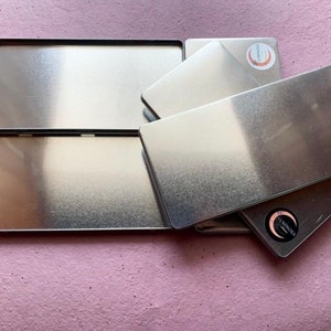May include: Five silver metal tins stacked on top of each other. The tins have a subtle gradient from dark to light. Each tin has a circular logo with the text "LUNAROSA" in a white font.
