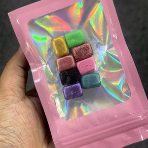 Könnte beinhalten: Ein pinkfarbener, wiederverschließbarer Beutel mit einem transparenten, holografischen Fenster, das neun kleine, rechteckige, farbige Wachsschmelzen enthält. Die Schmelzen sind in Gold-, Grün-, Rot-, Rosa-, Schwarz-, Lila- und Türkistönen gehalten. Der Beutel wird in einer Hand gehalten.