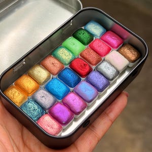 May include: A metal tin containing twenty-four rectangular watercolor pans with a shimmering finish in various colors including gold, blue, pink, green, and silver. The text "All Shimmers Mini Set of 24 pans" is printed on the lid of the tin.