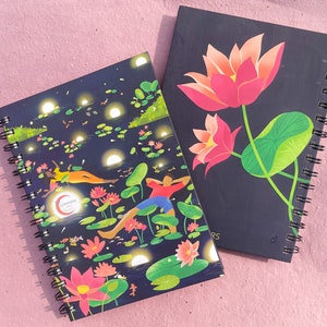 May include: Two spiral-bound notebooks. One features a dark blue cover with a nighttime scene of people relaxing among lily pads and glowing orbs. The other has a dark blue cover with pink lotus flowers and green leaves.