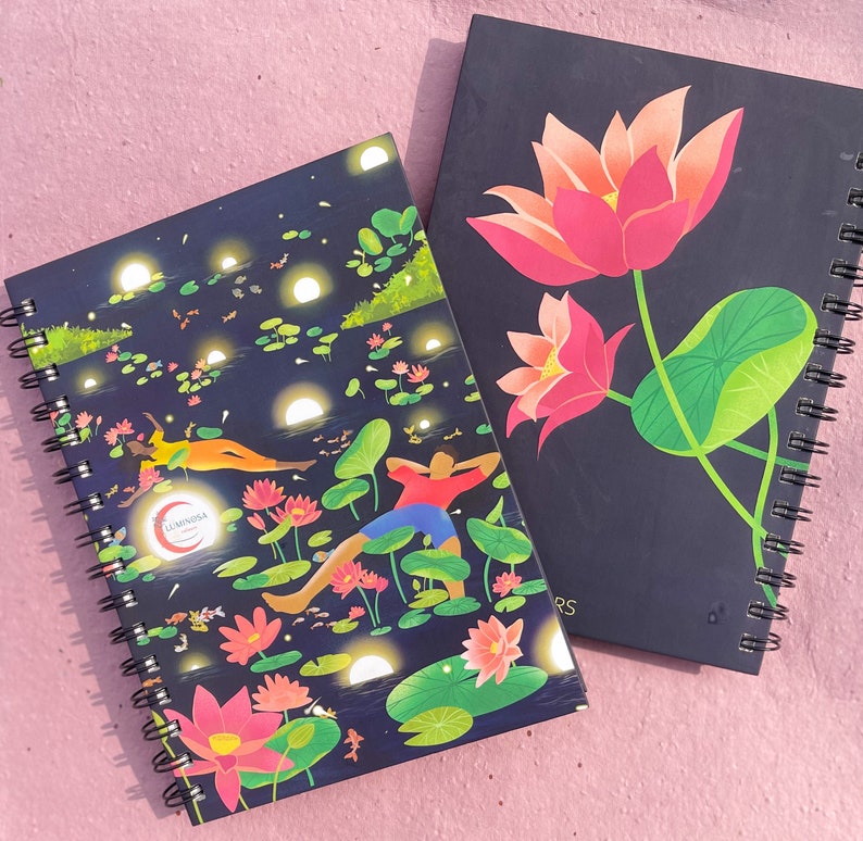 May include: Two spiral-bound notebooks. One features a dark blue cover with a nighttime scene of people relaxing among lily pads and glowing orbs. The other has a dark blue cover with pink lotus flowers and green leaves.