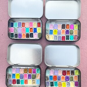 May include: Four open metal tins filled with watercolor paints. Each tin contains a grid of colorful, rectangular paint pans in shades of purple, pink, blue, green, yellow, orange, and red. The tins are on a pink surface.
