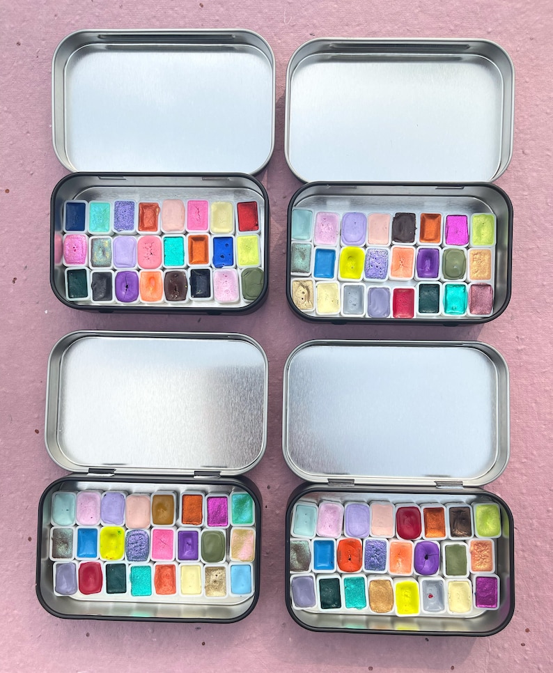 May include: Four open metal tins filled with watercolor paints. Each tin contains a grid of colorful, rectangular paint pans in shades of purple, pink, blue, green, yellow, orange, and red. The tins are on a pink surface.