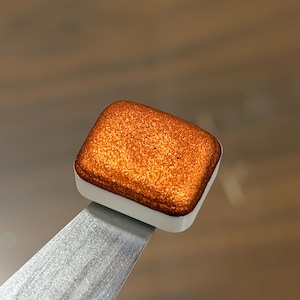 May include: A square watercolor paint pan featuring a vibrant, metallic copper pigment with a fine glittery texture. The paint is contained within a small, white plastic rectangular housing, which is held by a flat, silver-toned metal spatula tool.