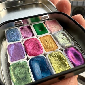 May include: A metal tin filled with 12 watercolor paint pans. The pans are arranged in a grid pattern and contain a variety of colors, including green, blue, purple, gold, and pink. The paint is iridescent and has a metallic sheen.
