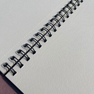 May include: Open sketchbook with blank white pages and a black spiral binding. The pages have a textured surface, and the binding is made of black metal loops. The sketchbook is open, revealing the clean, unused pages.