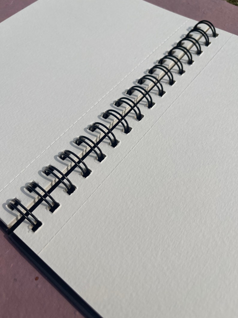 May include: Open sketchbook with blank white pages and a black spiral binding. The pages have a textured surface, and the binding is made of black metal loops. The sketchbook is open, revealing the clean, unused pages.