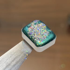 May include: A rectangular watercolor paint pan with a white plastic base. The paint surface features a deep metallic emerald green border surrounding a central cluster of dense, multi-colored holographic glitter particles that reflect light.