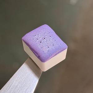 May include: A rectangular watercolor paint pan with a vibrant purple pigment featuring a textured, slightly porous surface. The paint is contained within a white plastic casing and is held by a pair of silver metal tweezers for display.