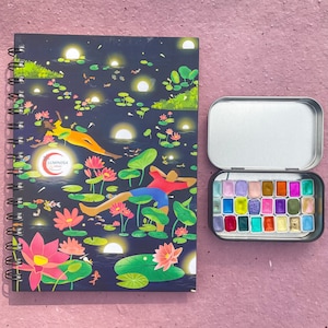 May include: A spiral-bound notebook with a dark blue cover featuring an illustration of people floating on lily pads with lotus flowers. Next to it is an open metal tin containing a set of watercolor paints in various colors.