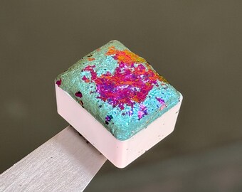 May include: A square watercolour paint pan with a light pink plastic base. The top surface features a textured, iridescent pigment with a vibrant colour shift, displaying patches of metallic teal, bright orange, and deep magenta throughout the surface.