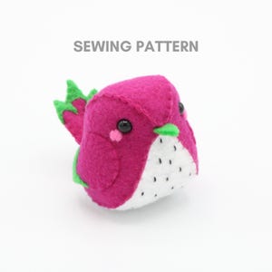 May include: A handmade felt dragon fruit plushie in shades of pink, white, and green. The plushie has black button eyes and a white belly with black seed details. The text "SEWING PATTERN" is above the plushie.