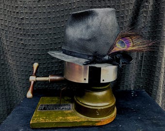 Puede incluir: Un sombrero negro con una pluma de pavo real en una máquina de estirar sombreros vintage. La máquina está hecha de metal y madera y tiene una manivela.