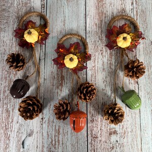 Könnte beinhalten: Drei herbstliche Hängedekorationen. Jede hat eine geflochtene Schlaufe, einen Blättercluster, einen kleinen gelben Kürbis und hängende Tannenzapfen, Eicheln und eine Glocke. Der Hintergrund ist eine verwitterte, hellfarbige Holzoberfläche.