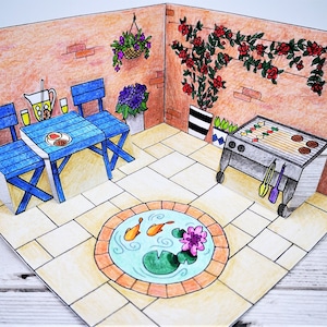 Könnte beinhalten: Eine Pop-up-Karte, die eine Terrasse mit einem blauen Tisch und Stühlen, einem Grill, einem kleinen Teich mit zwei orangefarbenen Fischen und einer Backsteinmauer mit einer roten Blütenranke zeigt.