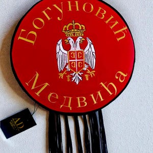 Puede incluir: Un emblema redondo rojo con letras doradas y un escudo de águila bicéfala. El emblema tiene el texto "Богуновић Медвѣђа" y una etiqueta que dice "AD Andrej Design". Flecos negros cuelgan en la parte inferior.
