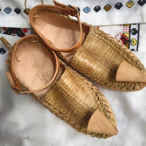 May include: A pair of woven leather sandals with a pointed toe and a strap that buckles at the ankle. The sandals are light brown and have a handwritten inscription on the inside.