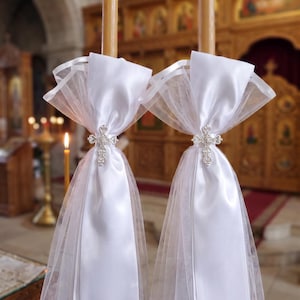 May include: Two tall, light brown taper candles decorated with white satin ribbon and sheer white tulle fabric. Each candle features a silver-toned metal cross ornament centred on the ribbon, creating a layered, decorative appearance.