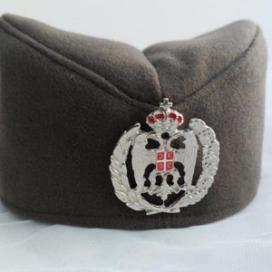 Puede incluir: Un gorro militar gris oscuro con un emblema plateado que presenta una corona, un águila y un escudo con detalles rojos y blancos. El gorro está hecho de un material suave, similar al fieltro.