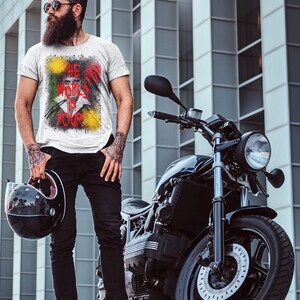 May include: A man with a beard and sunglasses is standing next to a black motorcycle. He is wearing a white t-shirt with a colourful graphic that says "The World Is Your".