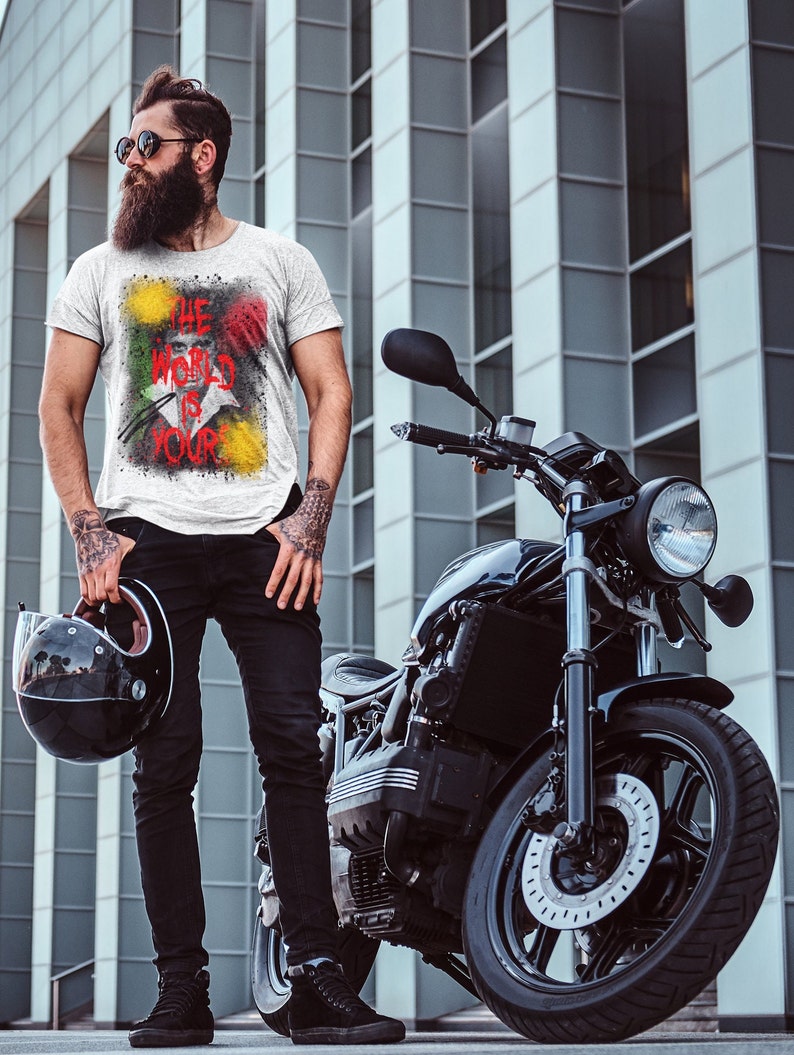 May include: A man with a beard and sunglasses is standing next to a black motorcycle. He is wearing a white t-shirt with a colourful graphic that says "The World Is Your".