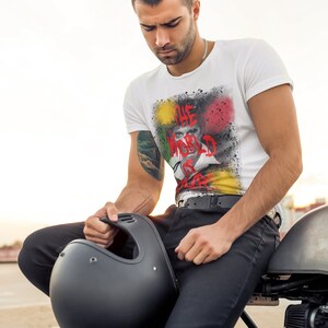May include: A man wearing a white t-shirt with a colourful graphic print that says "The World Is Play". He is sitting on a motorbike and holding a black helmet.