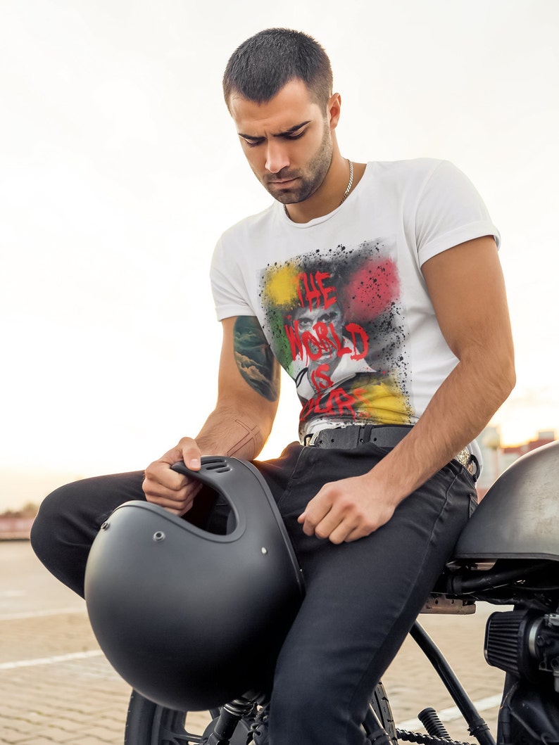 May include: A man wearing a white t-shirt with a colourful graphic print that says "The World Is Play". He is sitting on a motorbike and holding a black helmet.