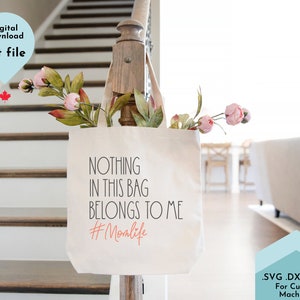 Puede incluir: Una bolsa de tela blanca con el texto "NOTHING IN THIS BAG BELONGS TO ME #Momlife" impreso en negro y naranja. La bolsa está colgada de una barandilla de madera con flores rosas en el fondo.