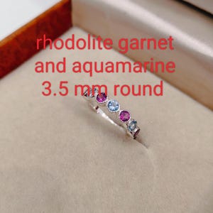 May include: A silver ring with alternating round rhodolite garnet and aquamarine gemstones. The ring features a series of vibrant pink and light blue stones. Text on the image reads "rhodolite garnet and aquamarine 3.5 mm round."