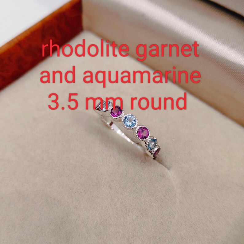 May include: A silver ring with alternating round rhodolite garnet and aquamarine gemstones. The ring features a series of vibrant pink and light blue stones. Text on the image reads "rhodolite garnet and aquamarine 3.5 mm round."