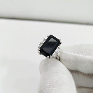May include: A silver ring featuring a large, rectangular black gemstone as the centerpiece. The ring is accented with smaller, rectangular white gemstones on the sides. The ring is held by a gloved hand against a white background.