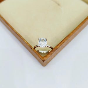 May include: A silver ring featuring a large, clear, cushion-cut gemstone. The ring is set in a simple, classic style and is presented in an open wooden jewellery box with a beige interior. The ring is centrally positioned.