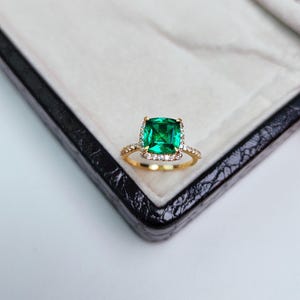 May include: A gold ring featuring a large, square-cut emerald center stone surrounded by a halo of small, clear gemstones. The ring is set on a delicate gold band. The ring is displayed in a black jewelry box with a white interior.