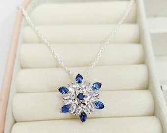 May include: A silver necklace with a snowflake pendant. The pendant features a central dark blue stone surrounded by white and blue teardrop-shaped stones. The necklace is displayed in a pink jewelry box.