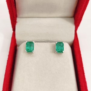 Puede incluir: Pendientes de botón con esmeraldas de talla esmeralda presentados en un joyero rojo. Los pendientes presentan gemas verdes vibrantes engastadas en una montura de plata u oro blanco. El interior de la caja es de color blanquecino.