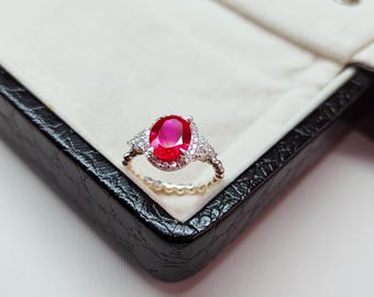 Puede incluir: Anillo de plata con una gran gema roja brillante de corte ovalado como pieza central. El anillo está engastado con gemas claras más pequeñas a ambos lados de la piedra principal. La banda tiene un diseño delicado y texturizado, presentado en una caja de joyería negra.