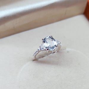 Op de afbeelding: Een zilverkleurige ring met een grote, ovaal geslepen, lichtblauwe middensteen die door vier pootjes wordt vastgehouden. De ringband is aan weerszijden bezet met drie kleine, heldere, rond geslepen steentjes voor een verfijnde uitstraling.