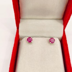 May include: A pair of round, deep red gemstone stud earrings set in silver-toned metal prongs. The earrings are displayed on a light-coloured, padded insert inside a bright red, open jewellery box with a white interior lining.