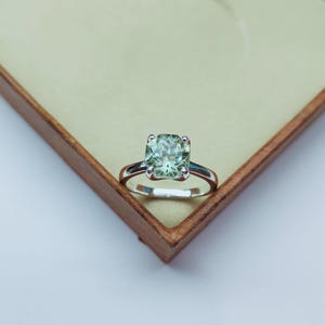 May include: A silver ring featuring a large, square-cut, pale green gemstone. The ring has a simple, classic design and is presented in a wooden jewellery box with a light beige interior. The ring is a piece of jewellery.