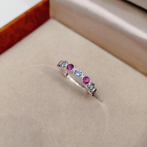 May include: A silver ring with a row of alternating round gemstones in shades of blue and magenta. The ring is set against a neutral background, highlighting the colorful stones.