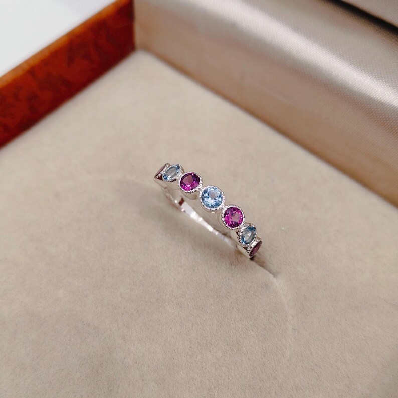 May include: A silver ring with a row of alternating round gemstones in shades of blue and magenta. The ring is set against a neutral background, highlighting the colorful stones.