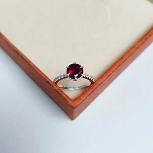 Könnte beinhalten: Ein silberner Ring mit einem großen, runden, tiefroten Edelstein als Mittelpunkt. Das Band ist mit kleineren, klaren Edelsteinen besetzt. Der Ring wird in einer hölzernen Schmuckschatulle mit beigefarbenem Innenraum präsentiert.