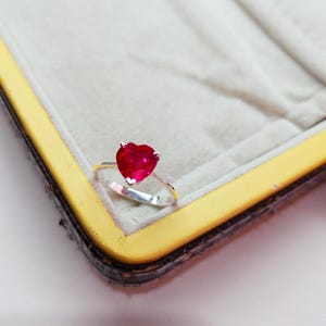 Op de afbeelding: Een zilveren ring met een hartvormige, helderrode edelsteen. De ring wordt gepresenteerd tegen een lichtgrijze achtergrond in een goudomrande sieradendoos, waarbij het ontwerp en de kleur van de ring worden benadrukt.