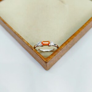 May include: A silver ring with an orange rectangular gemstone at the center, flanked by two smaller white gemstones. The ring is set against a light-colored background within a wooden box.