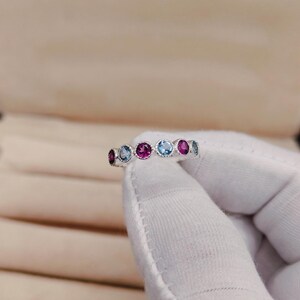 May include: A silver ring featuring a series of alternating gemstones. The ring showcases a pattern of round-cut gems in shades of pink, blue, and purple. The gemstones are set in a delicate, repeating design, creating a colorful and elegant piece of jewelry.